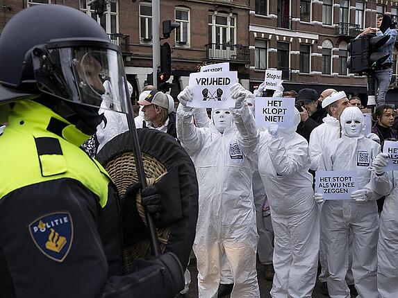 Menschen in Schutzanzügen und Masken halten Plakate hoch, während ein Bereitschaftspolizist im Vordergrund steht. Foto: Peter Dejong/AP/dpa